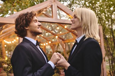 A charming outdoor setting features a young couple in elegant suits, holding hands and gazing lovingly at each other. Surrounded by greenery and soft, glowing lights, their bond shines brightly. clipart