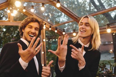 Two young men, dressed in formal attire, display their hands joyfully under a canopy of glowing lights in a greenhouse. Their laughter and genuine connection create a lively atmosphere. clipart