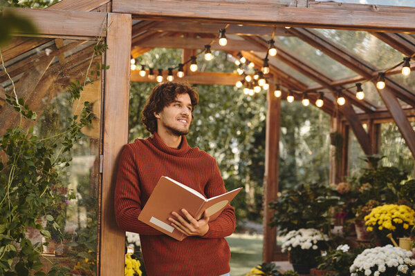 Surrounded by colorful flowers and soft lighting, a young man in a cozy sweater deeply engages with his book, creating a scene of tranquility in the greenhouse.