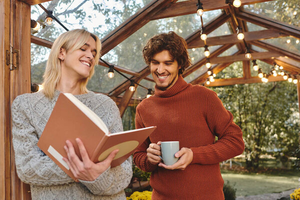Young loving gay couple smile as they engage with a book in a charming outdoor space adorned with lights. The atmosphere radiates warmth and connection as they enjoy each others company.