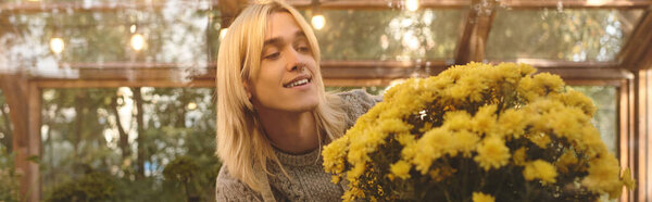 A young, handsome man tends to a large bouquet of bright yellow flowers inside a serene greenhouse. The setting is filled with natural light and greenery, creating a peaceful atmosphere.