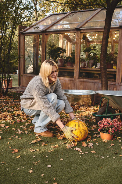 Молодой, красивый мужчина с светлыми волосами с радостью ухаживает за тыквой в пышном саду, наполненном осенними листьями..