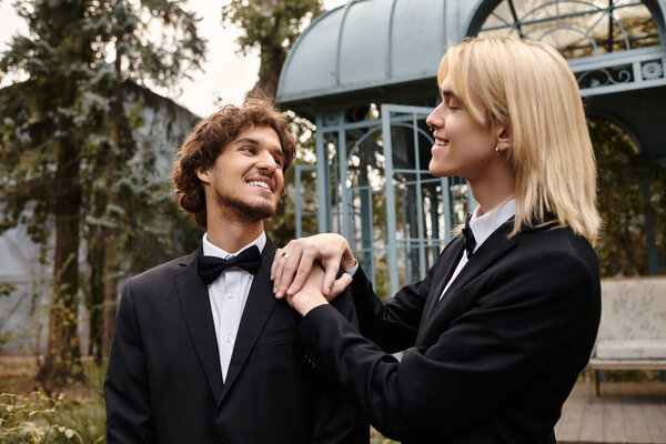 In a vibrant outdoor location, a young couple in elegant black suits share a joyful moment. Their expressions reflect happiness and affection, surrounded by lush greenery and a stunning backdrop.