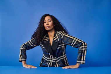 Kıvırcık saçlı şık bir kadın canlı mavi bir fon karşısında kendine güveniyor..