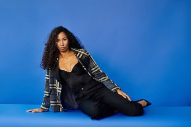 Kıvırcık saçlı kadın, mavi arka planda şık kıyafetler içinde şık bir poz verir..