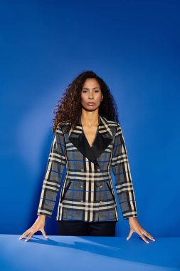 Ekose ceketli dengeli bir kadın zengin mavi bir arka plana karşı zarafet yayar..