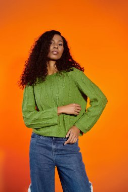 A confident woman in a green sweater poses gracefully against a vibrant orange background. clipart