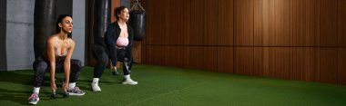 Kendini işine adamış iki kadın şık bir spor salonunda ağırlıklarla çömeliyor..