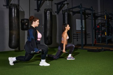 Two women perform lunges with weights in a spacious and well equipped gym environment. clipart