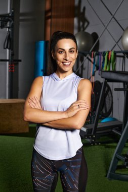 Kadın, iyi donanımlı bir spor salonuna özgüvenle poz veriyor. Kendini spora ve iyi olmaya adamış.