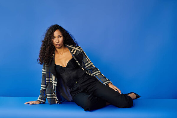 Кудрявая женщина застилает шикарную позу в стильной одежде на синем фоне.
