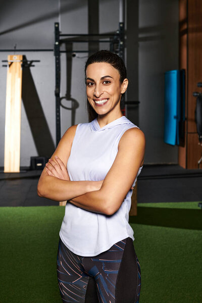 Уверенная женщина стоит со скрещенными руками в стильном спортзале, источая силу и мотивацию.