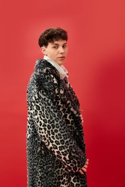 Adam çarpıcı bir leopar desenli ceket giyiyor, canlı kırmızı bir duvara karşı eşsiz bir stil sergiliyor..