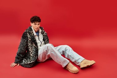 Cesur bir kırmızı arka plan karşısında oturan yakışıklı adam, eşsiz moda seçimleri ve özel yetenekleri sergiliyor..