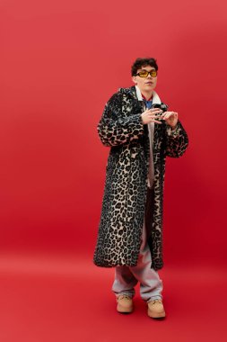 Çekici bir adam, leopar desenli ceketli ve gözlüklü canlı kırmızı bir arka plana karşı duruyor..