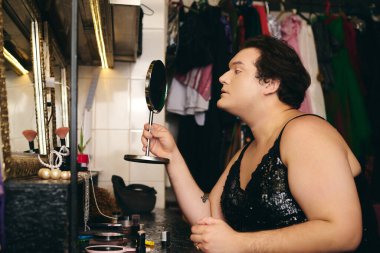 Bir travesti, ışıltılı bir soyunma odasında aynanın önünde, parlamaya hazır bir şekilde karmaşık bir makyaj yapar..