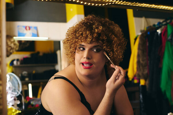 In a lively dressing room, a drag queen applies makeup, preparing for a festive show.