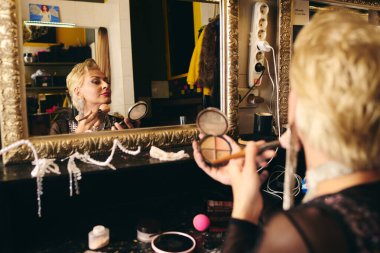 In a vibrant dressing room, a drag queen applies makeup, admiring their reflection. clipart