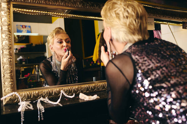 A skilled drag queen puts on bold lipstick, admiring their reflection in an ornate mirror.