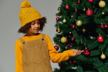 Sarı kazaklı ve şapkalı genç kız, Aralık 2025 'te bir Noel ağacına süs eşyası ekliyor.