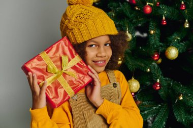 A little one beams with excitement, cradling a festive gift next to a sparkling tree. clipart