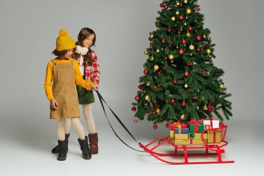 İki küçük kız Noel eğlencesinin tadını çıkarıyorlar, süslü bir ağacın yanında canlı bir kızak çekiyorlar..