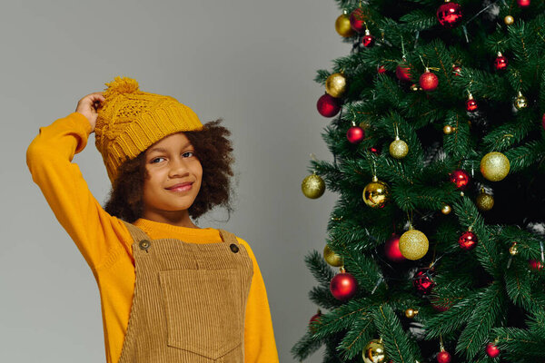 Веселая девушка в уютном желтом наряде рядом с праздничной, украшенной орнаментом елкой.