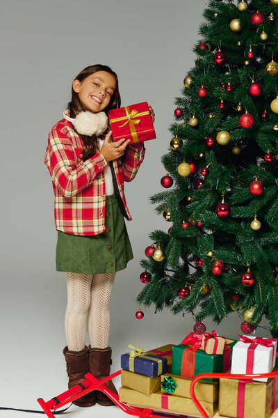 Маленькая девочка в праздничном наряде улыбается, держа подарок возле украшенной елки.