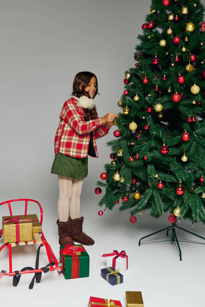 Девушка счастливо добавляет украшения к рождественской елке среди красочных подарков в праздничной обстановке.