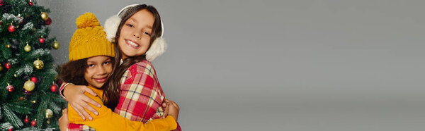 Две маленькие девочки счастливо обнимаются рядом с праздничной елкой, излучая радость и тепло.