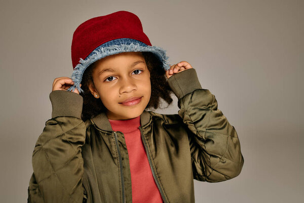 A cheerful little girl wears a red bucket hat and an olive green jacket with a warm smile.