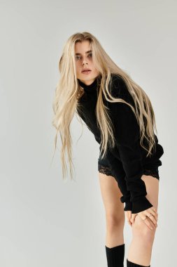 Genç kadın zarif pozlar veriyor. Uzun sarı saçlı, şık siyah elbiseli. Tarafsız bir arkaplana karşı..