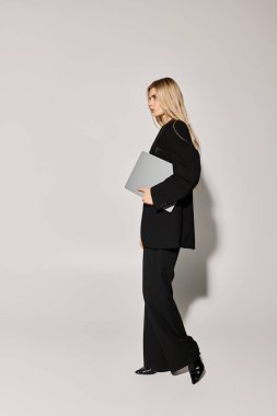 A stylish young woman with flowing blonde hair walks confidently, holding a laptop in a studio. clipart