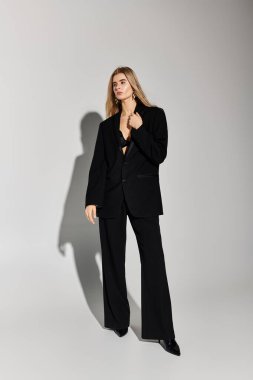 Uzun sarı saçlı, kendine güvenen genç bir kadın stüdyo arka planında siyah pozlar veriyor..