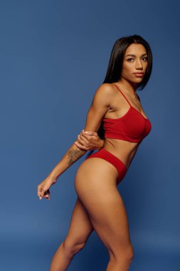 Esmer model, mavi arka planda kırmızı iç çamaşırlarıyla güvenle poz veriyor, çeşitliliği vurguluyor..