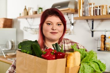 Young woman with vibrant hair happily holds a box of fresh vegetables in a warm kitchen. clipart