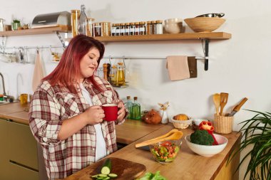 A beautiful plus size woman enjoys a warm drink amid fresh ingredients in her kitchen. clipart