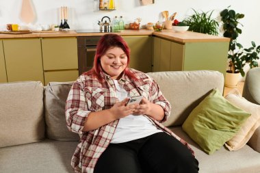 A beautiful young plus size woman smiles as she uses her phone in a stylish kitchen. clipart