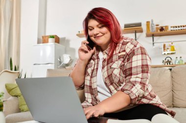 Genç ve cüsseli bir kadın bilgisayarını kullanırken neşeli bir telefon görüşmesi yapıyor.