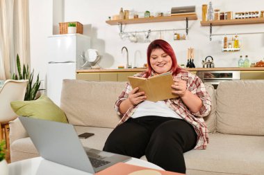 Güzel, şişman bir kadın evde rahat bir kanepede dinlenirken kitaptan zevk alıyor..