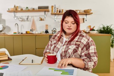 A young plus size woman studies graphs while enjoying a warm drink in a cozy kitchen clipart