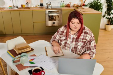 Artı boyda bir kadın, notları ve kahveyle birlikte şık bir mutfak masasında çalışmaya odaklanmış..