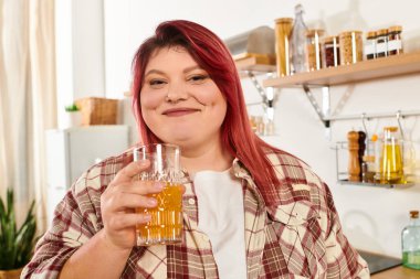 Joyful young woman with vibrant hair smiles while holding a drink in her stylish kitchen clipart