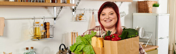 Веселая молодая женщина держит коробку свежих овощей, улыбаясь в своем ярком кухонном пространстве.
