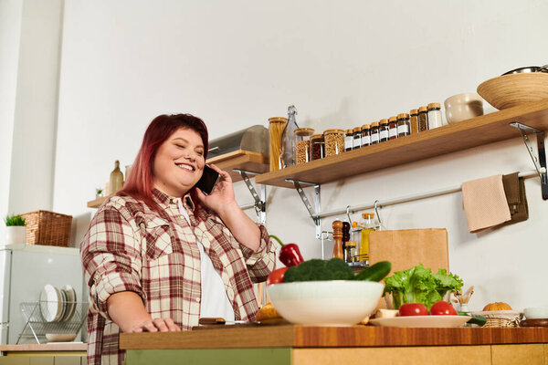 Молодая женщина улыбается и разговаривает по телефону, готовя свежие ингредиенты на стильной кухне.