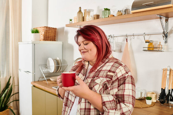 Joyful woman holds a red cup while savoring a warm drink in her stylish kitchen space.