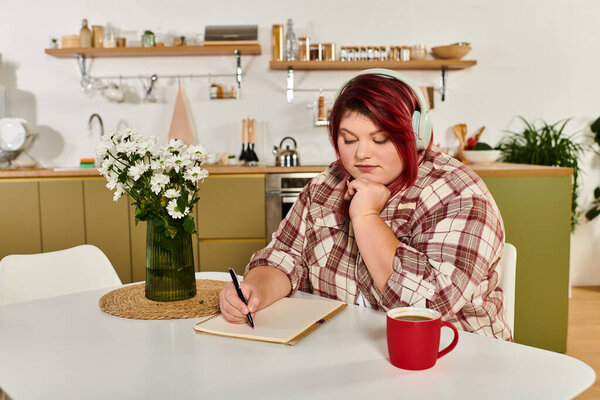 A young plus size woman enjoys writing while sipping coffee in a charming kitchen.