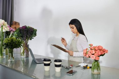 Brunette woman organizes flowers and takes notes in her vibrant flower shop, ready for customers. clipart