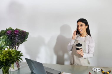Genç bir kadın çiçekçisini işletiyor, telefonda konuşuyor ve kahvenin tadını çıkarıyor..