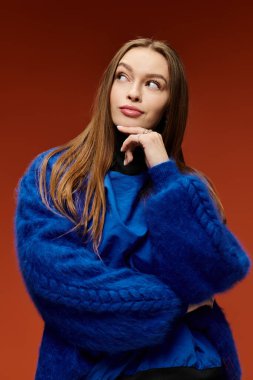 Parlak kıyafetli kendine güvenen bir kadın, uzun saçlarını sergileyerek, sıcak bir arkaplanda poz veriyor..